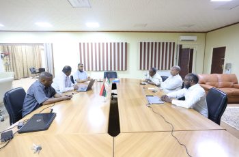 Six men sit around a long wooden conference table in a meeting room, with laptops and small flags at the center of the table.