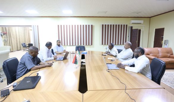 Six men sit around a long wooden conference table in a meeting room, with laptops and small flags at the center of the table.
