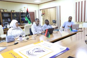 inbound1712292272493509759 - جامعة سبها | Sebha University People seated around a conference table in an office, with Libyan flags, laptops, and documents during a meeting.