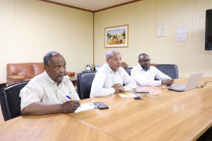 inbound2300396624433602898 - جامعة سبها | Sebha University Three men sit at a long conference table in a beige office, taking notes with a laptop nearby on the table.