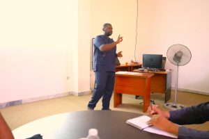 Male healthcare professional in navy scrubs giving a talk in a classroom, gesturing with his hands near a wooden desk with a laptop and papers nearby.