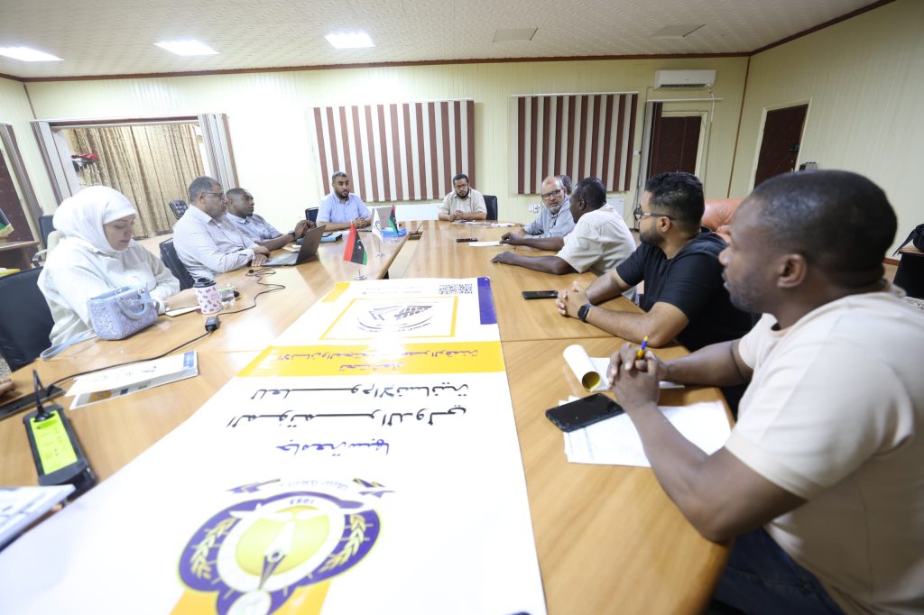 inbound438033717824845974 - جامعة سبها | Sebha University People seated around a long conference table discussing, with flags and posters on the table in a meeting room