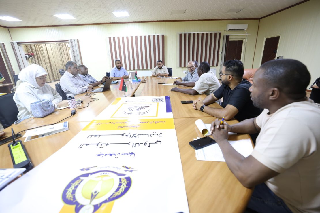 People seated around a long conference table discussing, with flags and posters on the table in a meeting room