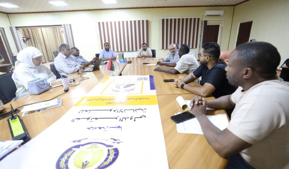 People seated around a long conference table discussing, with flags and posters on the table in a meeting room