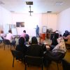 Instructor presenting to a group of adults in a classroom seminar with a wall screen and whiteboard at the front.