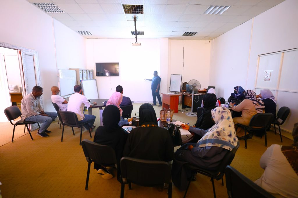 inbound4557199579059315725 - جامعة سبها | Sebha University Instructor presenting to a group of adults in a classroom seminar with a wall screen and whiteboard at the front.