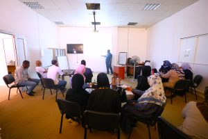 Instructor presenting to a group of adults in a classroom seminar with a wall screen and whiteboard at the front.