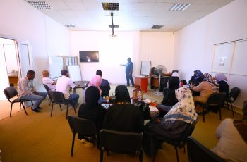 Instructor presenting to a group of adults in a classroom seminar with a wall screen and whiteboard at the front.