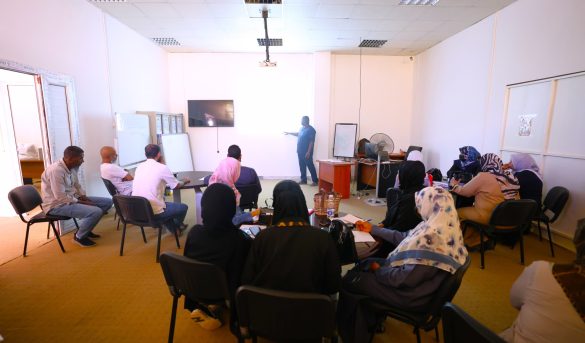 Instructor presenting to a group of adults in a classroom seminar with a wall screen and whiteboard at the front.
