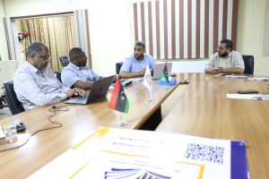 inbound5925623054072952155 - جامعة سبها | Sebha University Four men sit around a wooden conference table in a meeting room, laptops open and Libyan flags on the desk in front of them.