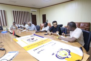inbound7955547787827612052 - جامعة سبها | Sebha University Group of men seated around a long conference table reviewing documents, with a large yellow poster and small flags on the table on the foreground.