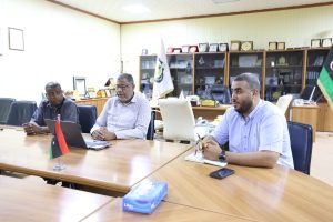inbound7991139476118946034 - جامعة سبها | Sebha University Three men sit at a conference table in an office, with flags and plaques in the background, appearing to discuss or review documents.