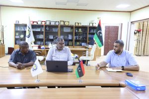 inbound8854747316592088884 - جامعة سبها | Sebha University Three men sit at a wooden conference table in a formal office, with a laptop open and Libyan flags in view.