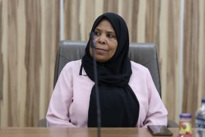 0011409D-722E-41D7-903A-E592CC628BBC - جامعة سبها | Sebha University Woman wearing a black hijab and light pink blazer sits at a conference table, looking to her left with a thoughtful expression.