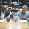 Two men in a formal office sign documents at a wooden table, with three small flags (Palestine, an institutional flag, and a Libyan-style flag) displayed in front of them and awards visible in the background.