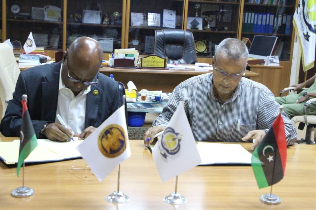 Two men in a formal office sign documents at a wooden table, with three small flags (Palestine, an institutional flag, and a Libyan-style flag) displayed in front of them and awards visible in the background.