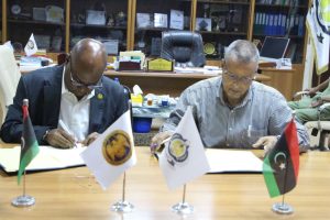 050B7EF4-E3A8-41A8-9DCB-E3FCDB04920F - جامعة سبها | Sebha University Two men in a formal office sign documents at a wooden table, with three small flags (Palestine, an institutional flag, and a Libyan-style flag) displayed in front of them and awards visible in the background.