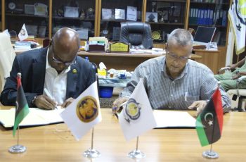 Two men in a formal office sign documents at a wooden table, with three small flags (Palestine, an institutional flag, and a Libyan-style flag) displayed in front of them and awards visible in the background.