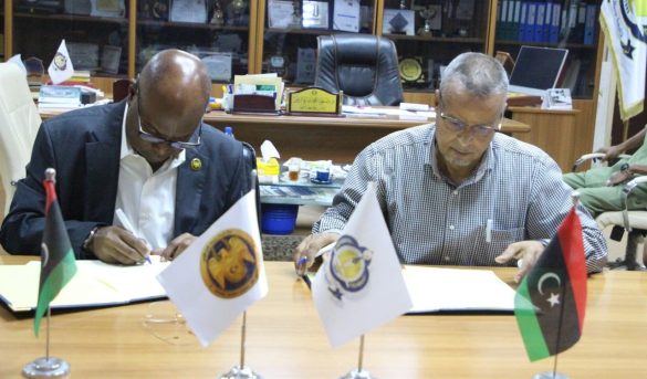 050B7EF4-E3A8-41A8-9DCB-E3FCDB04920F - جامعة سبها | Sebha University Two men in a formal office sign documents at a wooden table, with three small flags (Palestine, an institutional flag, and a Libyan-style flag) displayed in front of them and awards visible in the background.