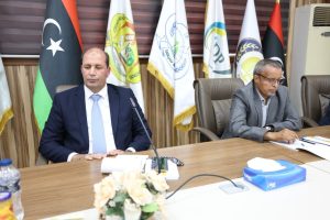 086FA7B3-5B7F-4149-945B-5A1E1CBCF8F6 - جامعة سبها | Sebha University Two men in suits sit at a conference table during a formal meeting, with flags in the background.
