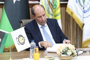 08848579-11B6-4DCB-8616-832D64C85529 - جامعة سبها | Sebha University Man in a dark suit and light blue tie signs documents at a conference table, with flags and a flower arrangement in the foreground at a formal meeting.