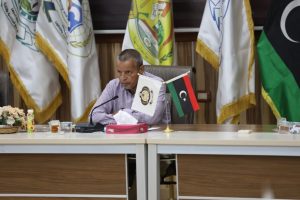 Man in a striped shirt sits at a conference table with a microphone, tissue box, and beverages, with Libyan and organizational flags in the background.
