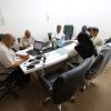 Seven people seated around a conference table in a meeting room with laptops, papers, and water bottles, collaborating at work.