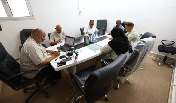 0C1C1879-074B-430B-A4C5-79A0217DC378 - جامعة سبها | Sebha University Seven people seated around a conference table in a meeting room with laptops, papers, and water bottles, collaborating at work.
