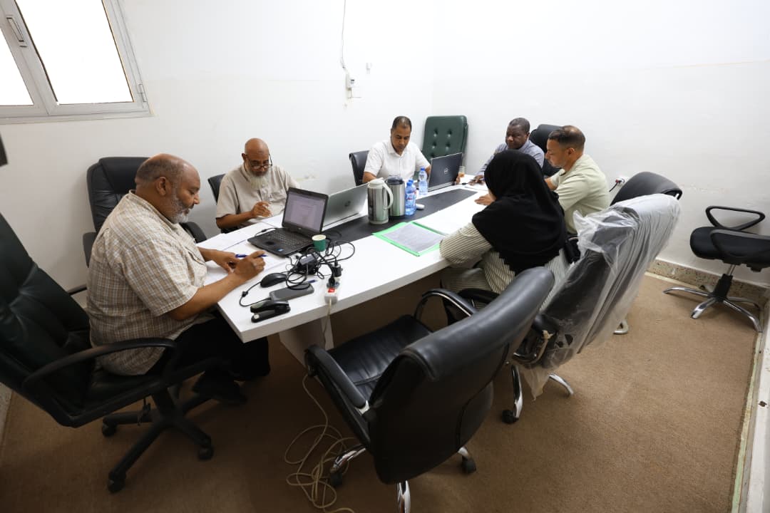 Seven people seated around a conference table in a meeting room with laptops, papers, and water bottles, collaborating at work.