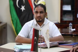Man in a white shirt sits at a desk in an office, hands folded, with Libyan flags and scattered documents in the background.