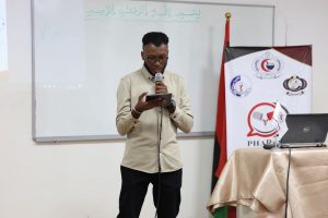 A man in a beige shirt speaks into a microphone while looking at a tablet on stage, in front of a whiteboard and banners.