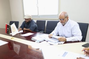 Two men sit at a glossy conference table signing documents; small flags stand nearby on the table in an office setting.