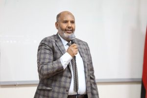Man in a brown plaid blazer speaks into a handheld microphone during a presentation.