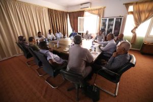 1CA34058-1295-46F0-B50E-FB8236CDE24C - جامعة سبها | Sebha University People seated around a large rectangular conference table in a meeting room, discussing with papers and bottled water on the table.