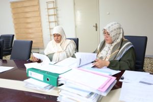 Two women in headscarves sit at a conference table reviewing documents and papers scattered across the desk.",