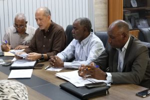 1E5BEBCD-4625-472B-9243-98257AB919FA - جامعة سبها | Sebha University Four men in a business meeting around a table, reviewing documents and taking notes.