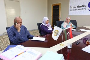 Three people sit at a university conference table; man on left talks on a smartphone while two women in hijabs review documents.