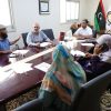 Six people sit around a glossy conference table in a formal meeting room, with papers, pens, and a tea glass on the table; Turkish flags and an emblem in the background.”