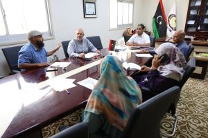 Six people sit around a glossy conference table in a formal meeting room, with papers, pens, and a tea glass on the table; Turkish flags and an emblem in the background.”