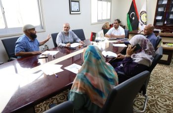 Six people sit around a glossy conference table in a formal meeting room, with papers, pens, and a tea glass on the table; Turkish flags and an emblem in the background.”