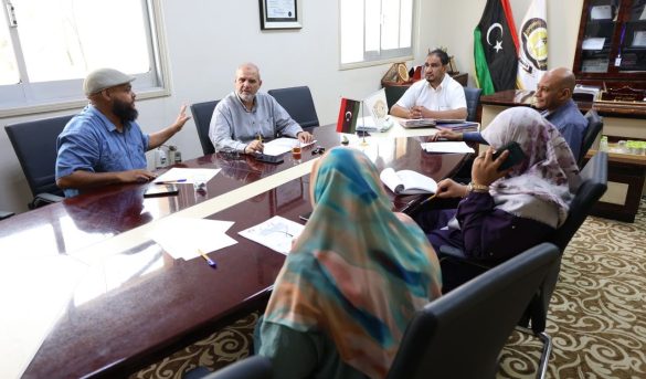 Six people sit around a glossy conference table in a formal meeting room, with papers, pens, and a tea glass on the table; Turkish flags and an emblem in the background.”