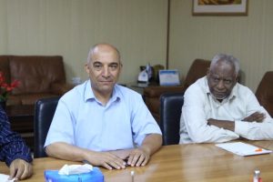 21005349-17C5-48D5-A27D-3268206C70BD - جامعة سبها | Sebha University Two men sit at a wooden conference table in a meeting room; one smiles in a light blue shirt, the other looks serious in a white shirt.