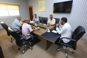 22F70E08-F5A6-48A5-AFA6-A971D5D70716 - جامعة سبها | Sebha University Six men sit around a rectangular conference table in a modern office, discussing with notebooks and coffee cups; a large TV and a framed map on the wall behind them.