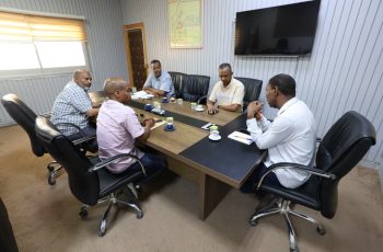 Six men sit around a rectangular conference table in a modern office, discussing with notebooks and coffee cups; a large TV and a framed map on the wall behind them.