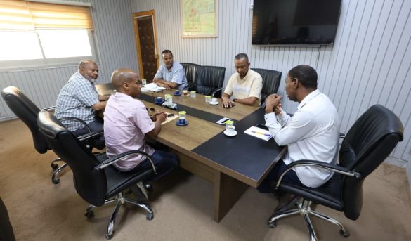 Six men sit around a rectangular conference table in a modern office, discussing with notebooks and coffee cups; a large TV and a framed map on the wall behind them.