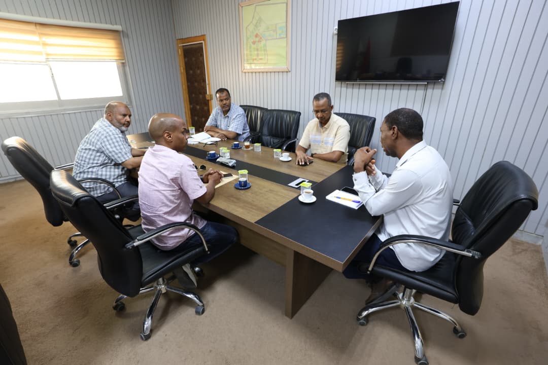 Six men sit around a rectangular conference table in a modern office, discussing with notebooks and coffee cups; a large TV and a framed map on the wall behind them.