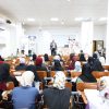 Male presenter on stage addressing a large audience of women in hijabs in a bright lecture hall