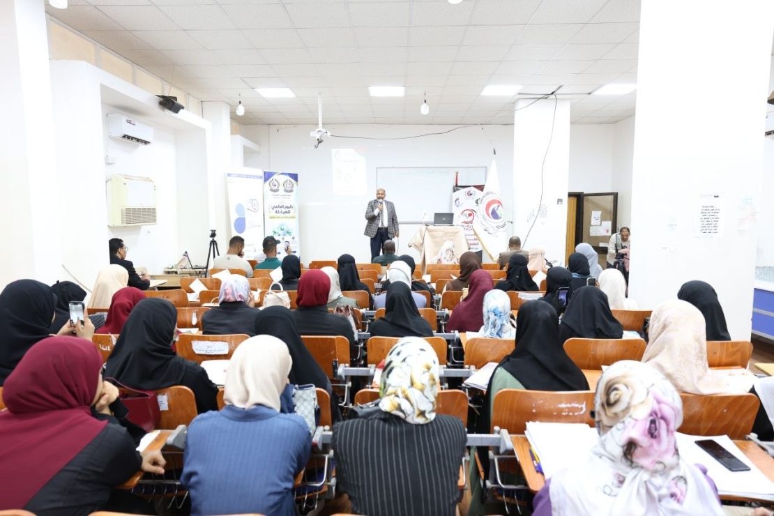 Male presenter on stage addressing a large audience of women in hijabs in a bright lecture hall