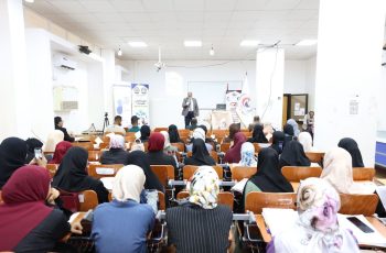 Male presenter on stage addressing a large audience of women in hijabs in a bright lecture hall