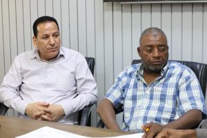 26EA6758-999C-4290-8E26-660F0FD9CF90 - جامعة سبها | Sebha University Two men sit at a conference table in an office; one wears a white plaid shirt, the other a blue plaid shirt, with papers on the table in front of them.